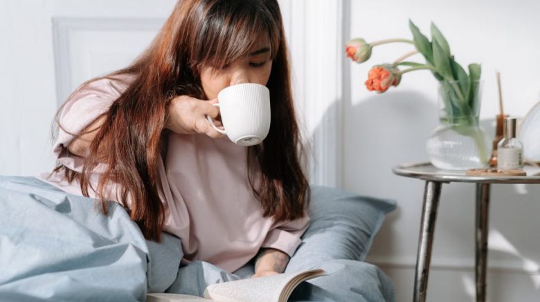 An Asian woman enjoying her morning with coffee and a book in a cozy bedroom setting.