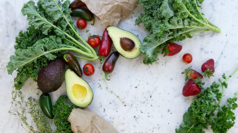 Top view of fresh avocados, kale, tomatoes, and peppers arranged on a marble surface.